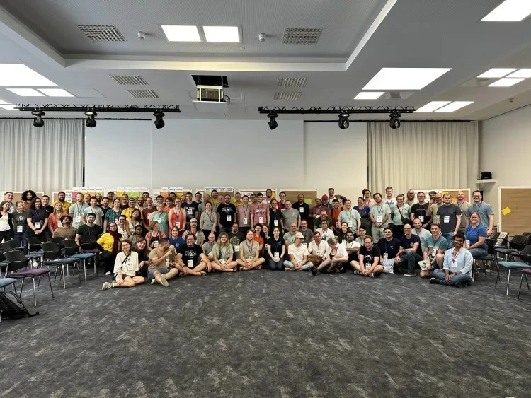 Gruppenfoto aller Teilnehmenden der SoCraTes Unconference 2025 in einem Konferenzraum, rund 100 Personen stehen und sitzen lächelnd in mehreren Reihen vor einer Pinnwand mit Session-Zetteln.